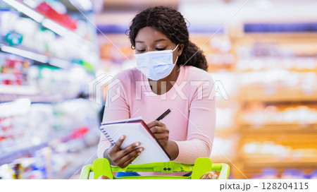African American woman in face mask doing shopping at supermarket, adding products to grocery list, writing in notebook. Black female customer purchasing goods at big mall African American woman in face mask doing shopping at supermarket, adding products to grocery list, writing in notebook. Black female customer purchasing goods at big mall 128204115