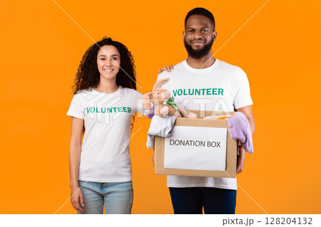 Couple Of Volunteers Posing With Donation Box Peeking Clothes For Charity Over Yellow Background, Smiling To Camera. Studio Shot. Volunteering Work, Contribution Concept Couple Of Volunteers Posing With Donation Box Peeking Clothes For Charity Over Yellow Background, Smiling To Camera. Studio Shot. Volunteering Work, Contribution Concept 128204132