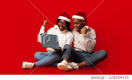 Christmas Offer. Excited black couple in santa hats celebrating success with laptop over orange background, cheerful african spouses sitting with computer and raising fists with joy, copy space Christmas Offer. Excited black couple in santa hats celebrating success with laptop over orange background, cheerful african spouses sitting with computer and raising fists with joy, copy space 128204133