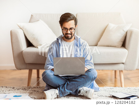 Remote Work Concept. Smiling Arab Freelancer Guy Using Laptop Computer At Home, Sitting On Floor In Living Room, Young Eastern Man Enjoying Working Remotely And Distant Job Opportunities, Copy Space 128204182
