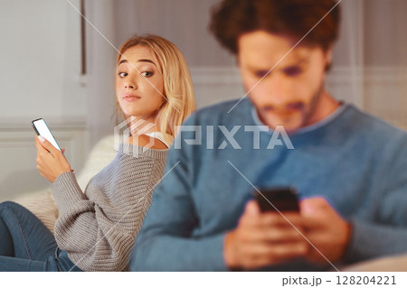 Cheating Signs. Girl Looking At Her Boyfriend Texting On Phone Suspecting Infidelity Sitting On Couch At Home. Selective Focus Cheating Signs. Girl Looking At Her Boyfriend Texting On Phone Suspecting Infidelity Sitting On Couch At Home. Selective Focus 128204221