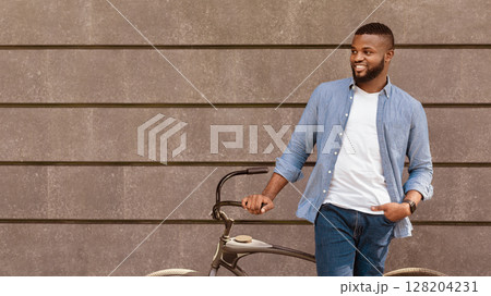Portrait of handsome black guy in casual clothes standing with his bike next to gray urban wall and looking away, panorama with copy space 128204231