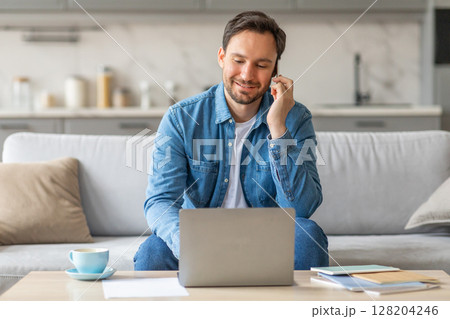 A man sits on a white couch in a living room and works on his laptop while speaking on a phone. He is wearing a blue jean shirt and smiling. On the table in front of him is a cup of coffee A man sits on a white couch in a living room and works on his laptop while speaking on a phone. He is wearing a blue jean shirt and smiling. On the table in front of him is a cup of coffee 128204246