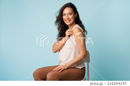 Coronavirus Vaccination. Happy Young Lady Showing Vaccinated Arm With Medical Adhesive Bandage Strip After Covid-19 Vaccine Injection Over Blue Studio Background. Covid Virus Immunization Campaign Coronavirus Vaccination. Happy Young Lady Showing Vaccinated Arm With Medical Adhesive Bandage Strip After Covid-19 Vaccine Injection Over Blue Studio Background. Covid Virus Immunization Campaign 128204247