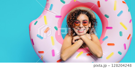 A young black woman is smiling happily as she poses inside a donut-shaped inflatable pool float. She is wearing sunglasses and a hat, and the background is a vibrant blue A young black woman is smiling happily as she poses inside a donut-shaped inflatable pool float. She is wearing sunglasses and a hat, and the background is a vibrant blue 128204248