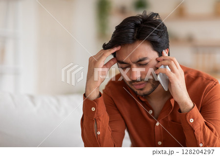 A young Arab man with a beard sits on a couch while talking on his phone. He appears stressed as he holds his forehead and frowns, copy space A young Arab man with a beard sits on a couch while talking on his phone. He appears stressed as he holds his forehead and frowns, copy space 128204297