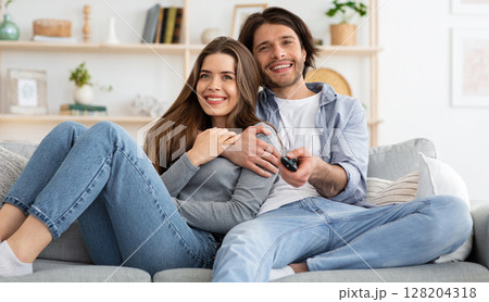 Joyful young couple watching TV together at home, embracing and sitting on couch. Cheerful man and woman switching channels on TV with remote, seeking nice movie for weekend, empty space 128204318