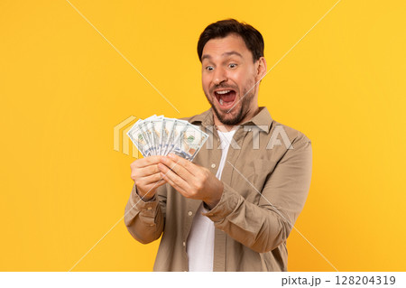 A man with a beard and brown shirt stands in front of a yellow background, smiling and holding a fan of dollar bills. His eyes are wide and he is clearly excited and surprised. A man with a beard and brown shirt stands in front of a yellow background, smiling and holding a fan of dollar bills. His eyes are wide and he is clearly excited and surprised. 128204319