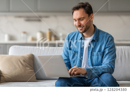 Man in a blue button-down shirt sits on a white couch in a living room. He is smiling and working on a laptop, which is resting on his lap. He appears to be focused on his work and enjoying his time. 128204380