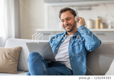 A man sits on a white couch in his living room, wearing a denim shirt, jeans, and headphones. He smiles as he looks at his laptop and adjusts his headphones with one hand. 128204383