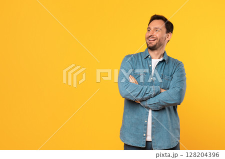 A man with a beard and short brown hair wears a blue denim shirt with a white undershirt. He stands in front of a yellow background, smiling, with his arms crossed. He is looking at copy space 128204396