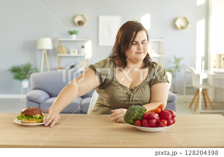 Plus size woman refusing eating hamburger as keeping to diet chooses plate with healthy vegetables 128204398