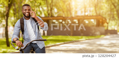 Pleasant Call. Cheerful Black Guy Talking On Cellphone While Riding Bicycle Outdoors, Enjoying Ride In City Park, Free Space 128204427