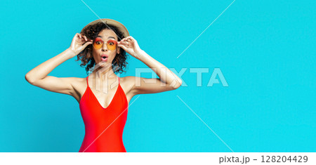 A young black woman with curly hair is wearing a red swimsuit, a straw hat, and sunglasses. She has her mouth open in surprise. She is standing in front of a solid turquoise background, copy space A young black woman with curly hair is wearing a red swimsuit, a straw hat, and sunglasses. She has her mouth open in surprise. She is standing in front of a solid turquoise background, copy space 128204429