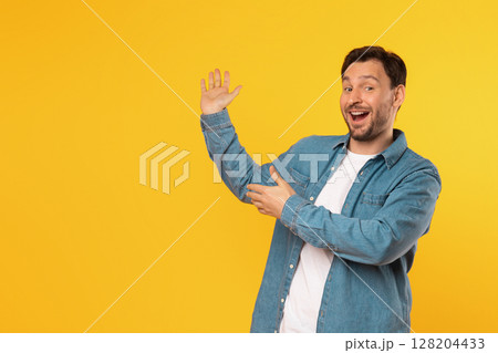A man wearing a denim shirt and a white t-shirt stands in front of a bright yellow background. He points at copy space and he is looking directly at the camera with a wide smile 128204433