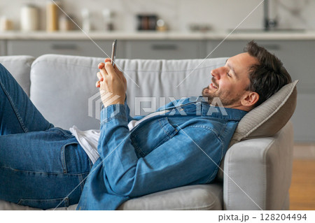 A man lies on a grey couch, propped up on a pillow, with his legs outstretched and his arms resting on the couch. He is using his smartphone with a smile on his face. 128204494