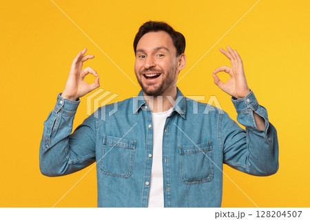 A smiling man wearing a blue denim shirt is standing against a bright yellow background. He is making the okay gesture with both hands. A smiling man wearing a blue denim shirt is standing against a bright yellow background. He is making the okay gesture with both hands. 128204507