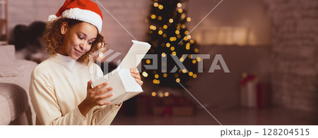 Christmas time. Cute afro girl looking inside gift box in front of xmas tree at home Christmas time. Cute afro girl looking inside gift box in front of xmas tree at home 128204515