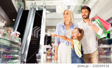 Family Shopping Concept. Little Girl Pointing Finger At Something Walking With Parents In Mall. Free Space For Text Family Shopping Concept. Little Girl Pointing Finger At Something Walking With Parents In Mall. Free Space For Text 128204516