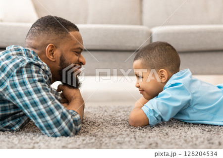 Happy African Father And Son Looking At One Another Lying On Floor At Home. Dad And Kid Bonding. Child And Daddy's Relationship And Trust Concept. Side View Portrait 128204534