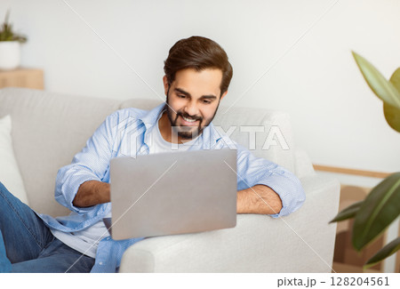 Online Job. Smiling Arab Freelancer Guy Using Laptop At Home, Handsome Eastern Man Sitting On Couch With Computer, Working Remotely And Enjoying Distance Business Opportunities, Free Space 128204561