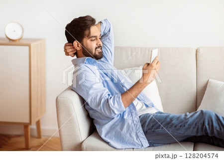 Millennial eastern guy resting on couch with smartphone, browsing internet. Young arab man relaxing with cellphone on sofa in living room, enjoying online communication, side view with free space Millennial eastern guy resting on couch with smartphone, browsing internet. Young arab man relaxing with cellphone on sofa in living room, enjoying online communication, side view with free space 128204574