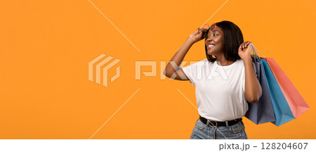 Joyful young black lady with sunglasses and shopping bags posing over orange studio background. Smiling african american woman removing glasses and holding purchases, looking at copy space 128204607