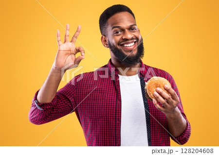 Cheerful African American Guy Posing With Burger Gesturing Okay Sign Smiling To Camera Standing On Yellow Studio Background. Black Man Approving Junk Food Taste 128204608