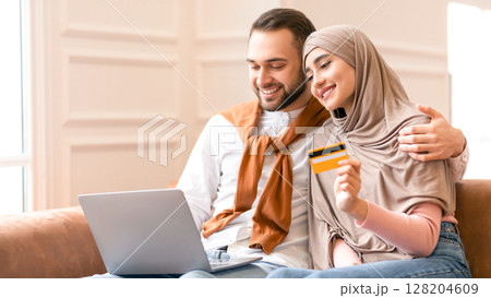 Muslim Family Of Two Shopping Online At Laptop Holding Credit Card Sitting On Couch At Home. Arab Couple Shops On Weekend. Internet Shop Sales And Discounts Concept. Selective Focus 128204609