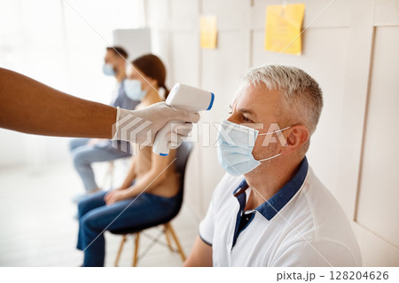Senior man in medical mask waiting for covid-19 vaccination at hospital, doctor checking his temperature with infrared thermometer. Immunization against coronavirus, population protection Senior man in medical mask waiting for covid-19 vaccination at hospital, doctor checking his temperature with infrared thermometer. Immunization against coronavirus, population protection 128204626