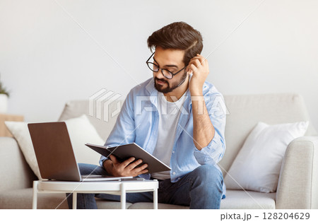 Young Arab Freelancer Guy Checking Working Schedule In Planner Notepad At Home. Millennial Eastern Man Sitting At Table With Laptop Computer, Enjoying Remote Work And Distance Job Opportunities 128204629