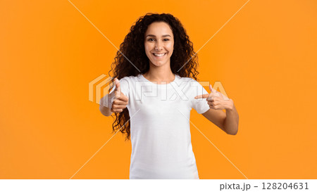 I'm Cool. Cheerful Young Woman In White T-Shirt Gesturing Thumbs Up Pointing Finger At Herself Standing On Yellow Background In Studio, Smiling To Camera. Choose Me Concept. Panorama 128204631