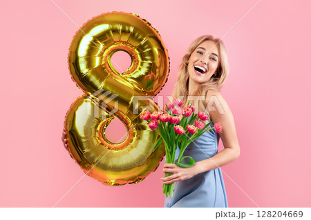 Cheerful millennial lady with bouquet of flowers and number eight balloon on spring holiday over pink studio background. Joyful young blonde celebrating Woman's Day. Festive concept 128204669