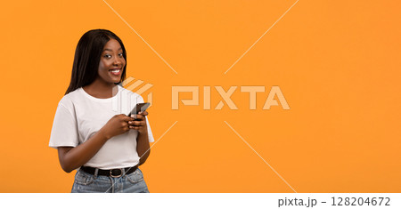 Beautiful young black lady in white t-shirt using smartphone over orange studio background, panorama with copy space. Cute african american woman chatting with friends, surfing on social media 128204672