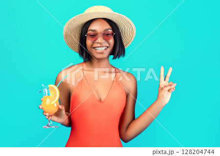 Positive black woman in bright swimsuit posing with tropical fruit cocktail, showing victory gesture on blue studio background. Beautiful African American lady holding exotic summer drink Positive black woman in bright swimsuit posing with tropical fruit cocktail, showing victory gesture on blue studio background. Beautiful African American lady holding exotic summer drink 128204744