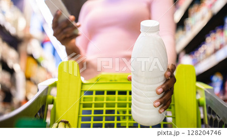 Young black woman checking her shopping list in smartphone app, putting bottle of milk into her cart at supermarket, selective focus. Low angle view. African American lady buying groceries at mall 128204746