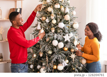 Home comfort, New Years decor and self-isolation. Young smiling african american husband and wife decorate Christmas tree with toys and balls in living room interior, side view, free space Home comfort, New Years decor and self-isolation. Young smiling african american husband and wife decorate Christmas tree with toys and balls in living room interior, side view, free space 128204781