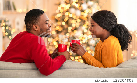 New year mood at home and romantic celebration together. Smiling millennial african american couple with cups with hot drink in interior of living room with Christmas tree and glowing garlands New year mood at home and romantic celebration together. Smiling millennial african american couple with cups with hot drink in interior of living room with Christmas tree and glowing garlands 128204803