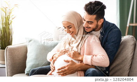 Happy islamic couple expecting baby, hugging on couch at home, cheerful muslim spouses relaxing together on sofa in living room, tenderly touching pregnant belly and smiling, copy space 128204804