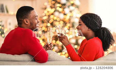 Christmas, New Year and comfort of home. Happy young african american couple in love clink glasses with champagne, sit on sofa with tree with garlands in modern living room interior, free space Christmas, New Year and comfort of home. Happy young african american couple in love clink glasses with champagne, sit on sofa with tree with garlands in modern living room interior, free space 128204806