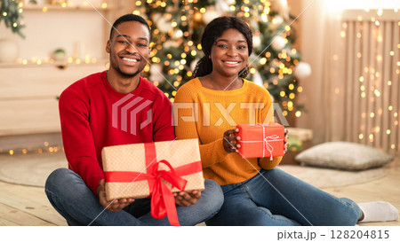 Gifts for family and New Years home decor. Millennial happy african american couple sit on floor and hold boxes with present in living room interior with Xmas tree and glowing garlands, free space Gifts for family and New Years home decor. Millennial happy african american couple sit on floor and hold boxes with present in living room interior with Xmas tree and glowing garlands, free space 128204815