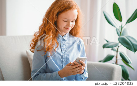 Digital Natives Concept. Positive smiling red-haired teenage kid using her cellphone, watching video online, sitting on the couch in living room or bedroom at home. Girl scrolling social media feed Digital Natives Concept. Positive smiling red-haired teenage kid using her cellphone, watching video online, sitting on the couch in living room or bedroom at home. Girl scrolling social media feed 128204832