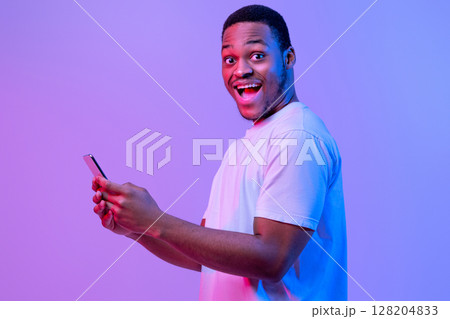 Mobile Offer. Portrait Of Excited African American Man Holding Smartphone, Cheerful Black Guy Enjoying Modern Technologies, Standing Under Neon Light Over Purple Studio Background, Copy Space 128204833