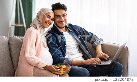 Pregnant Couple's Leisure. Happy Expectant Islamic Spouses Relaxing In Living Room, Young Arab Husband Spending Time With Laptop While His Muslim Wife In Hijab Enjoying Vegetable Salad, Free Space Pregnant Couple's Leisure. Happy Expectant Islamic Spouses Relaxing In Living Room, Young Arab Husband Spending Time With Laptop While His Muslim Wife In Hijab Enjoying Vegetable Salad, Free Space 128204878
