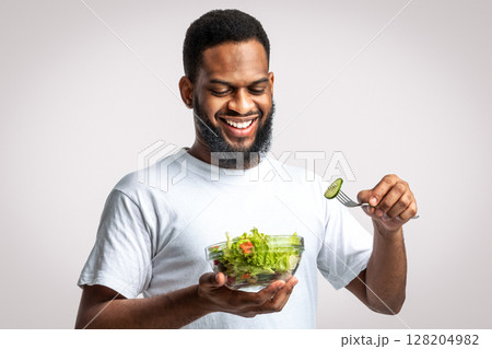 Healthy Eating. Happy Black Man Eats Fresh Vegetable Salad Slimming Standing Posing Over White Studio Background. Male Weight Loss, Nutrition And Dieting Concept Healthy Eating. Happy Black Man Eats Fresh Vegetable Salad Slimming Standing Posing Over White Studio Background. Male Weight Loss, Nutrition And Dieting Concept 128204982