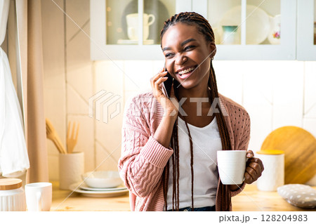 Cheerful black woman talking on mobile phone and drinking coffee in kitchen at home, positive black lady holding mug with hot drink and enjoying pleasant conversation with friend, free space 128204983