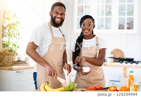 Happy Millennial African American Couple Wearing Aprons Cooking Healthy Lunch Together In Kitchen, Cheerful Black Spouses Enjoying Preparing Food At Home, Looking At Camera And Smiling, Free Space 128204994