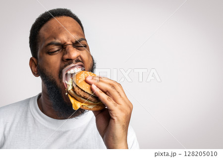 African Man Biting Burger Enjoying Unhealthy Tasty Junk Food Standing In Studio Over White Background. Overeating, Fast Food And Male Nutrition Concept. Funny Hungry Black Guy Eats Cheeseburger 128205010