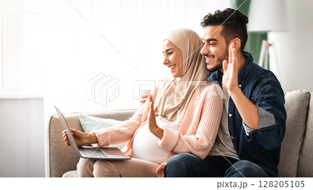 Cheerful Pregnant Muslim Couple Using Laptop At Home For Video Call, Happy Expectant Middle Eastern Spouses Talking To Family Or Friends Via Web Conference, Waving Hands At Computer Camera, Side View Cheerful Pregnant Muslim Couple Using Laptop At Home For Video Call, Happy Expectant Middle Eastern Spouses Talking To Family Or Friends Via Web Conference, Waving Hands At Computer Camera, Side View 128205105