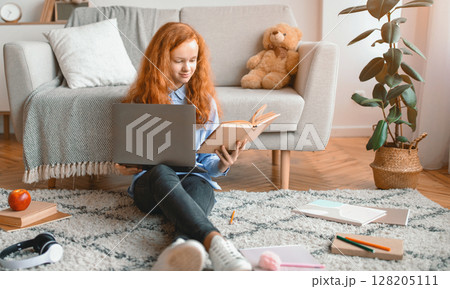Preparing For Exam. Smart smiling casual gen z girl sitting on the floor carpet at home in living room, leaning on couch, holding paper book and using laptop computer, doing homework 128205111